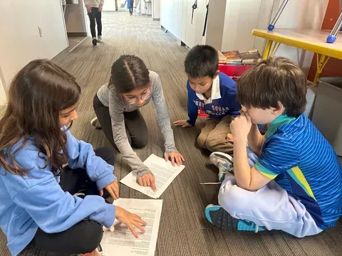four students seated on the ground work together to read a worksheet