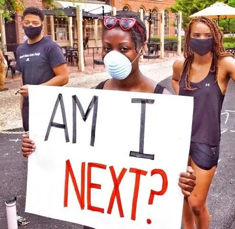 Hayes, wearing a medical face mask, holds up a sign that reads "Am I next?" Two other young people stand in the background.