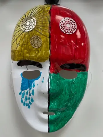 Close-up of a student mask divided into yellow, red, white, and green quadrants. They yellow quadrant has a flower design, and medallions are placed along the forehead. Beneath the right eye hole, a cloud of tears falls.