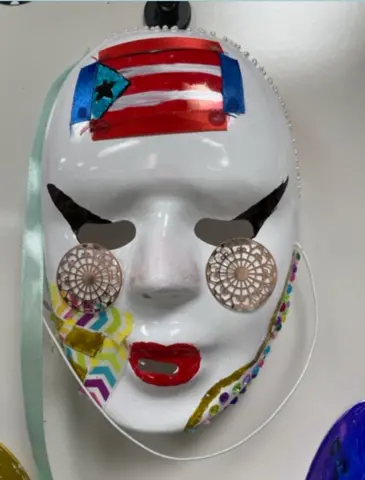 Close-up of a student mask with winged eyes, medallions under the eyes, red painted lips, jewels and chevrons along the jaw, and a Puerto Rican flag in the center of the forehead.