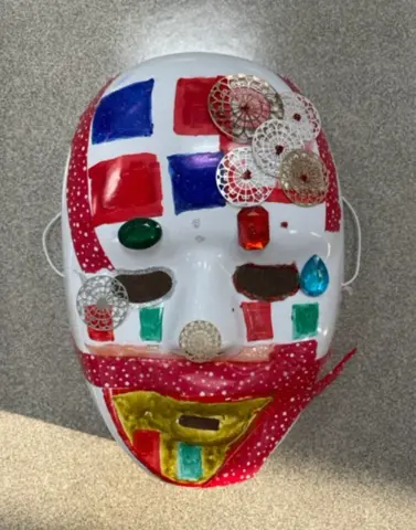 Close-up of a student mask with red and white tiles, jewels around the eyes, and a golden mouth surrounded by speckled red.