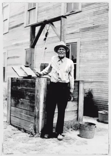 Nap McQueen, an elderly Black man poses in front of a well for a portrait. He is wearing a button down shirt, slacks, and a hat