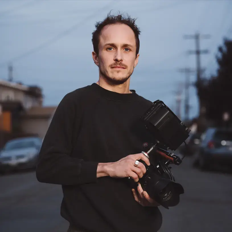 Headshot of Brandon Tauszik. He wears a black long-sleeved crewneck sweatshirt and holds a video camera.