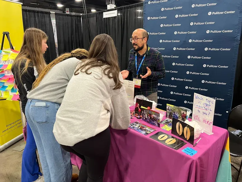 Fareed Mostoufi, the Pulitzer Center’s associate director of K-12 Education, stands at a Pulitzer Center-branded table in a conference exhibit hall, speaking with three students whose backs are facing the camera.