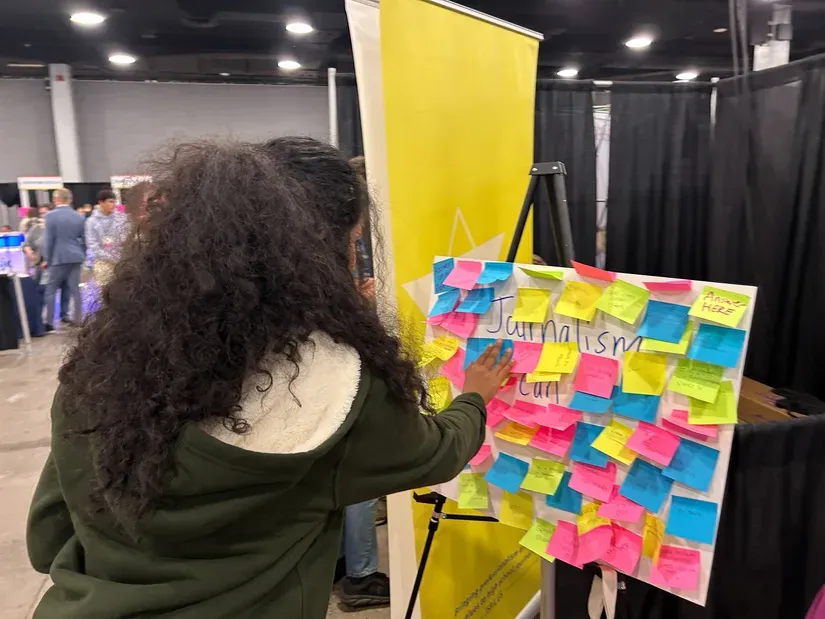 A student places a post-it note on a posterboard that poses the question "What can journalism do?" in a conference exhibit hall.