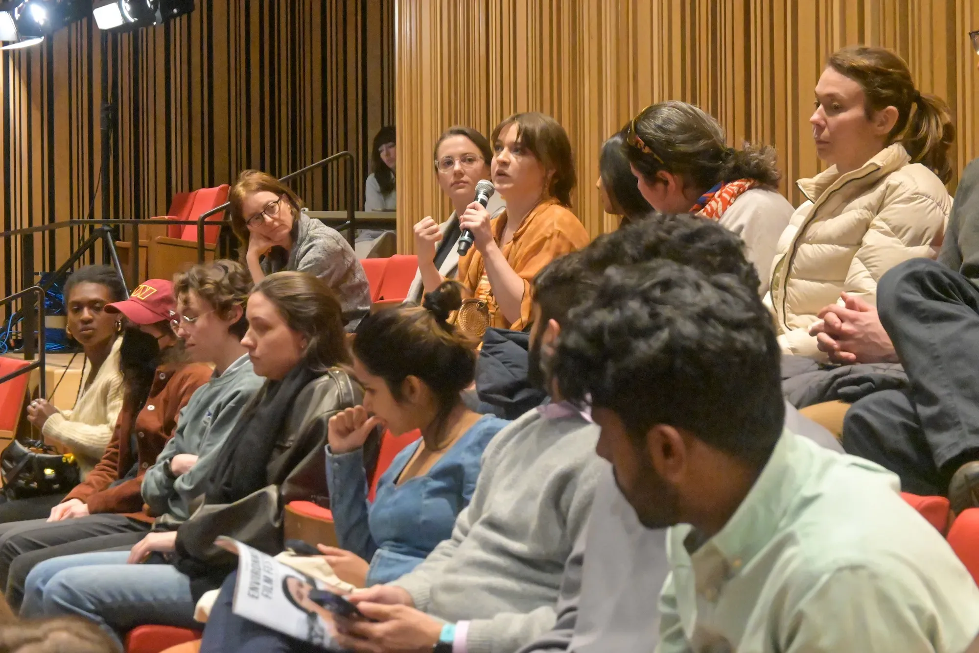 Woman in orange shirt sits in audience and asks a question 