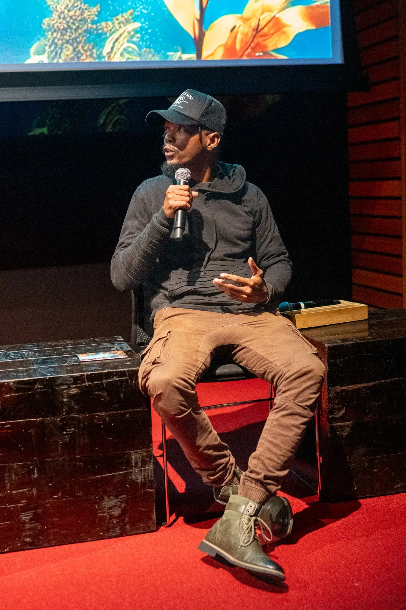 Man speaks into microphone in black zip up and hat. 