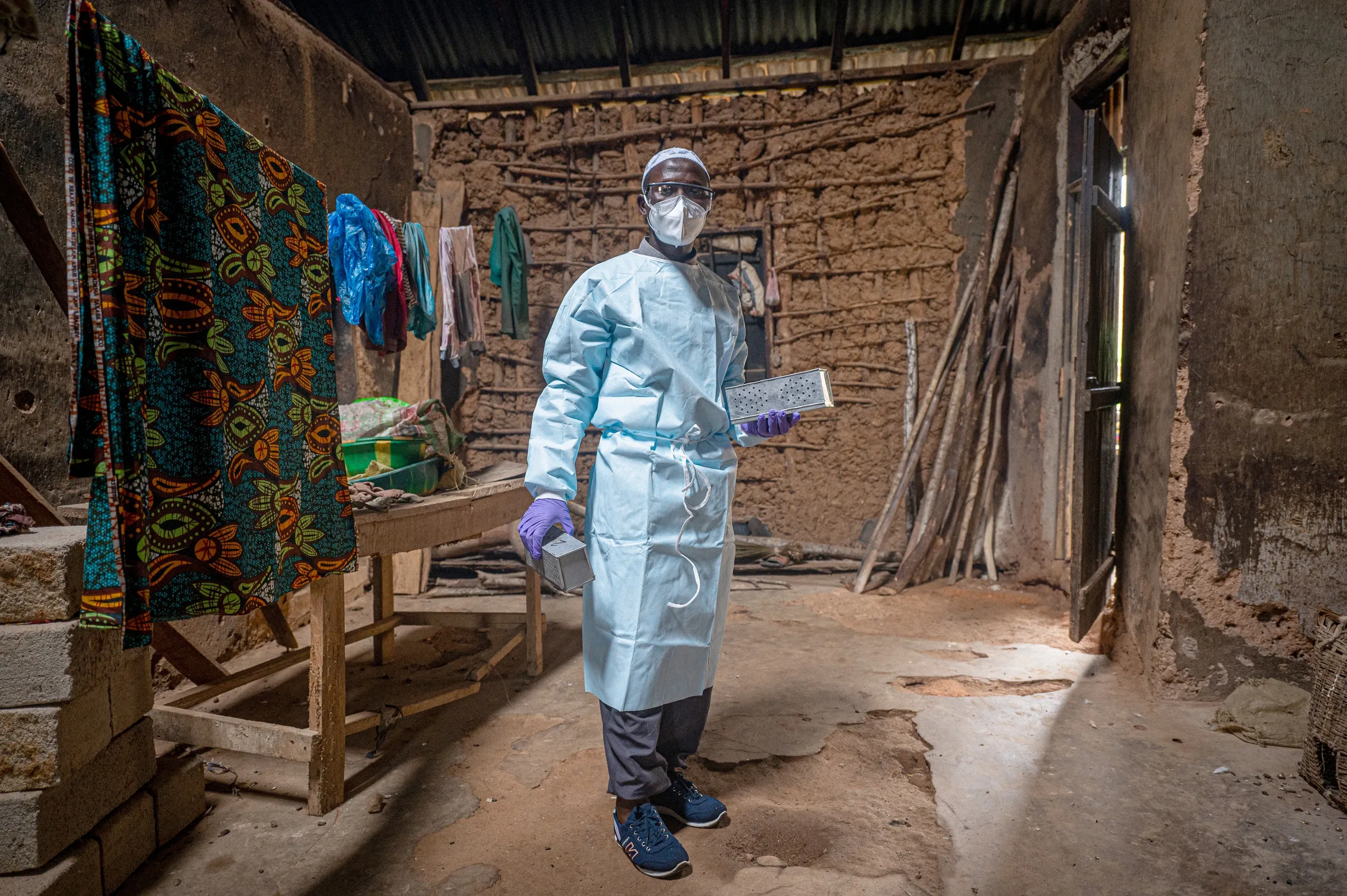 A medical professional in protective wear, gloves, mask and goggles holds two metal boxes as he stands in the middle of an building made from mud bricks