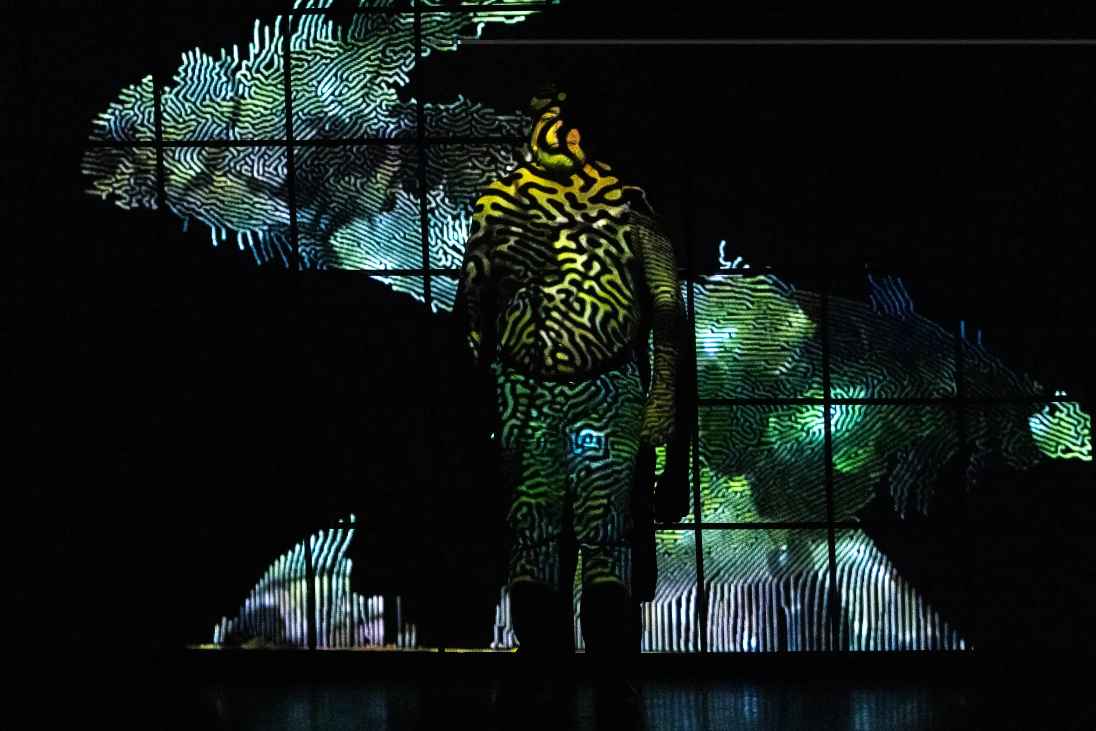 On a stage, a shirtless man is shrouded in shadow as parts of his abdomen are lit by projected graphic design dominated by green and black lines