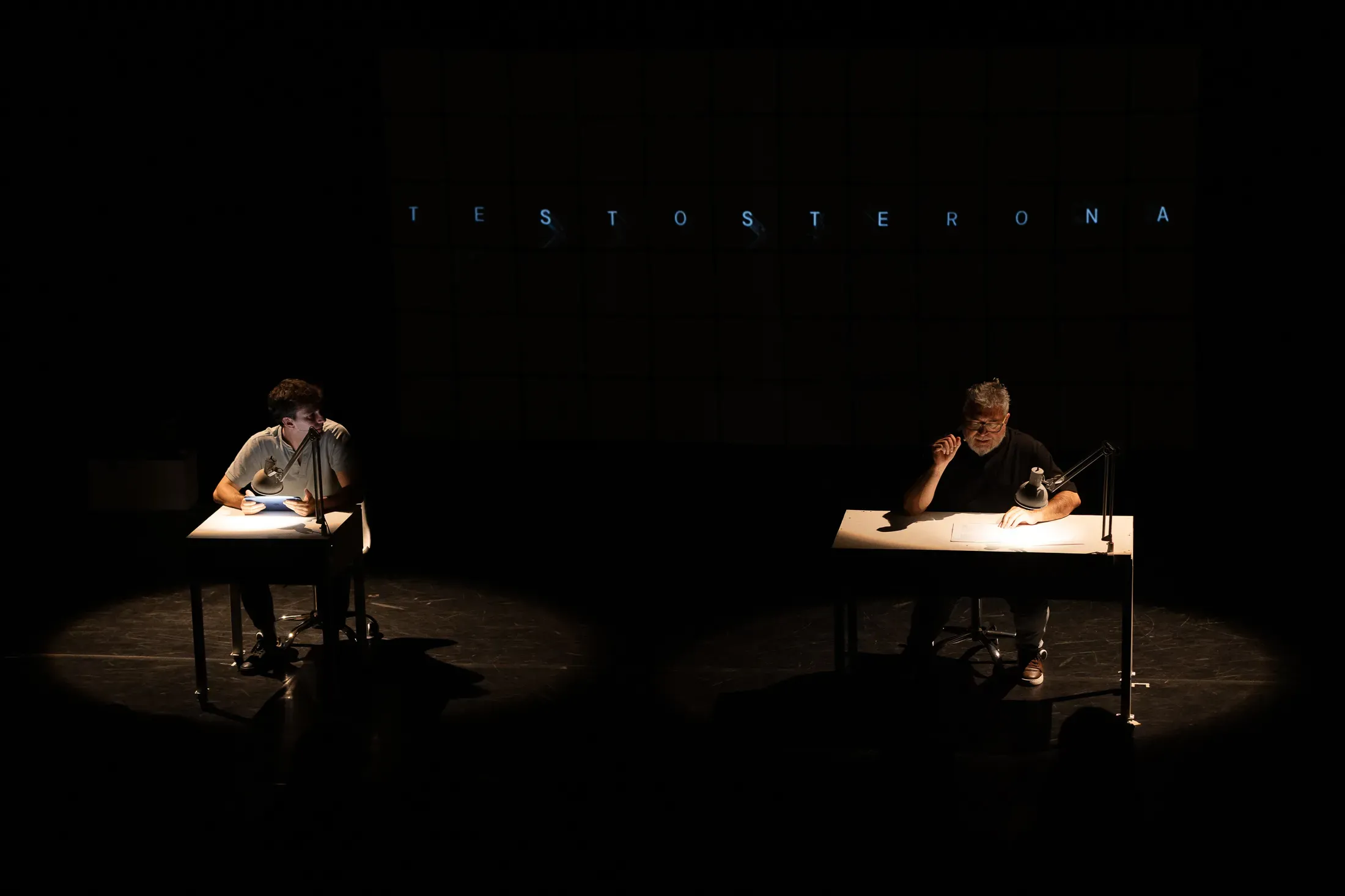 Two men on stage are separately lit as they sit at school desks