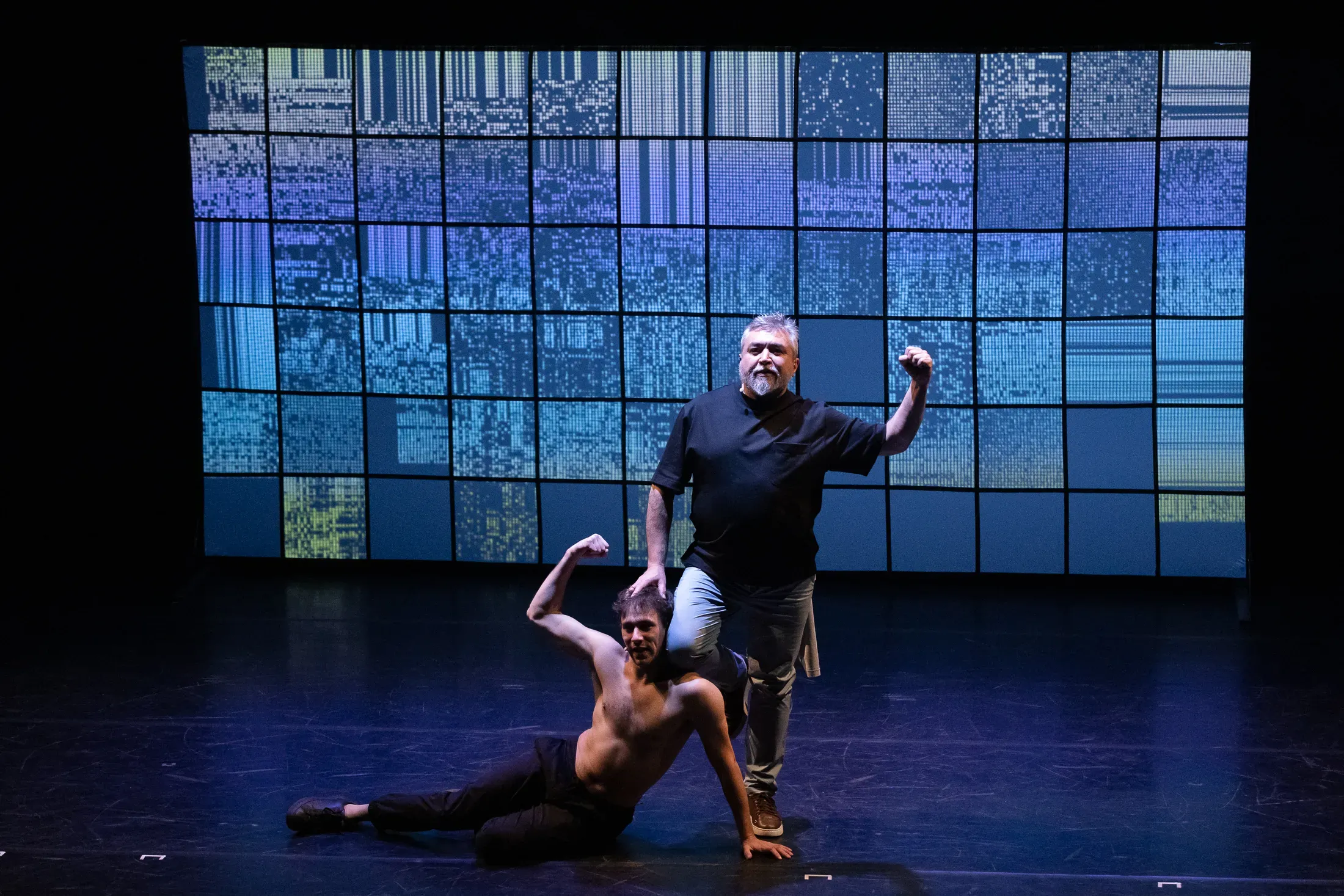 On a well lit stage, one man kneels at the feet of another, who raises a fist into the air.