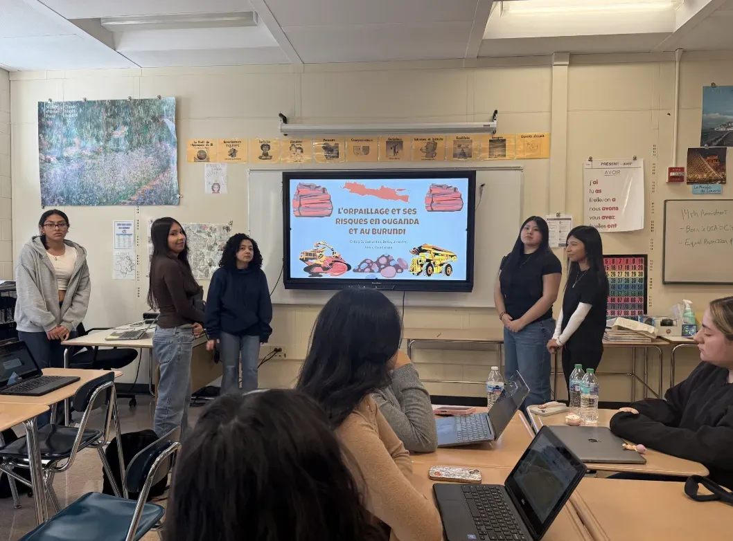 students stand at the front of the classroom beside screen to share a presentation 