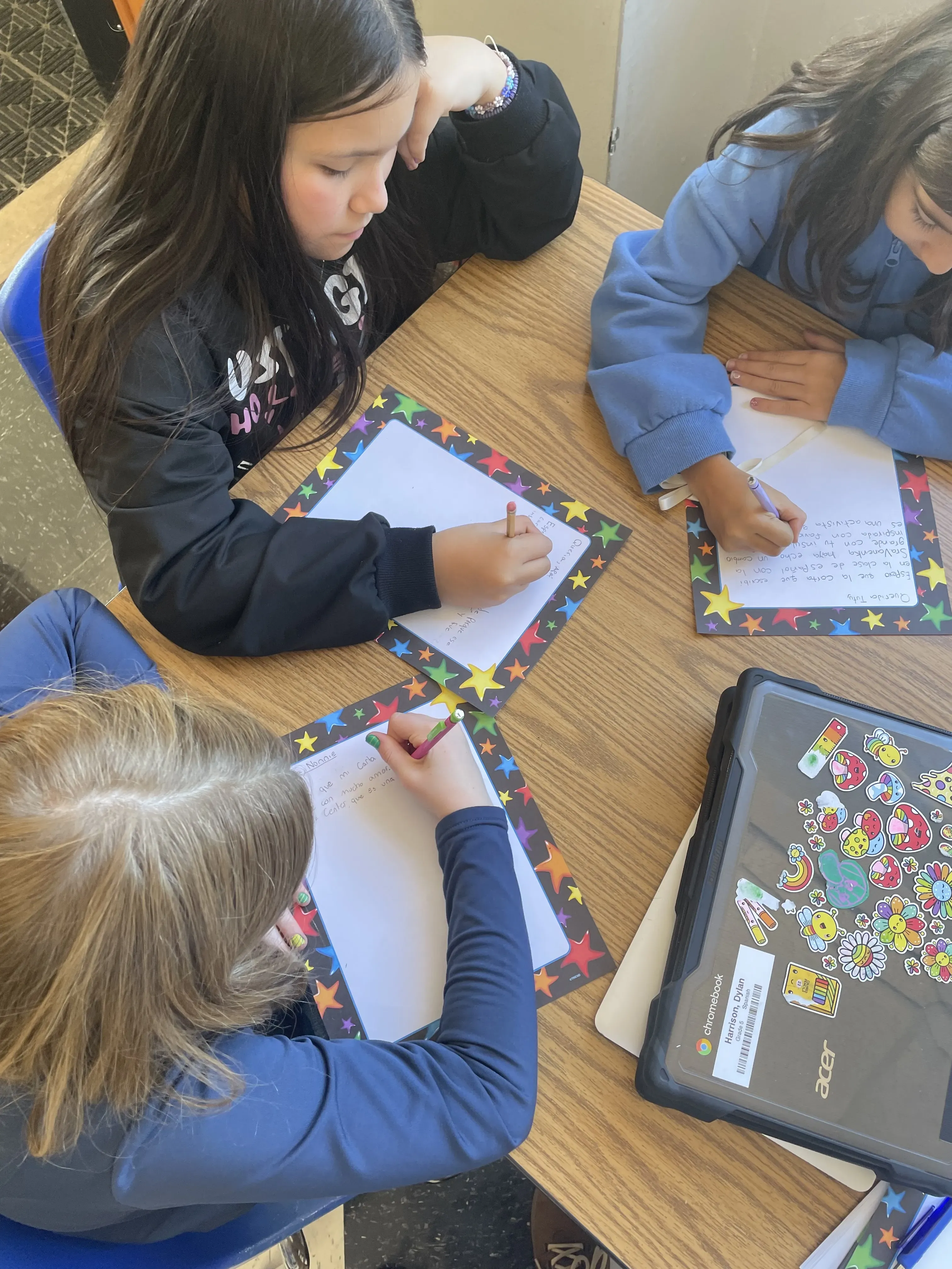 three students seated at table write letters on paper with colorful star border