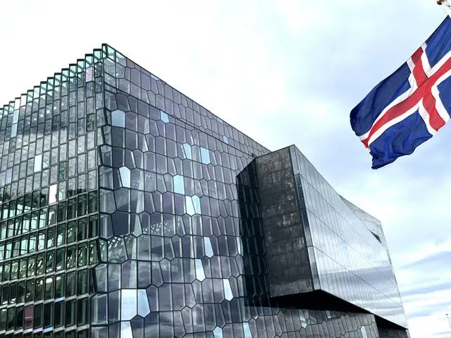 The Icelandic national flag waves outside a large building covered entirely with organically-shaped glass panels.