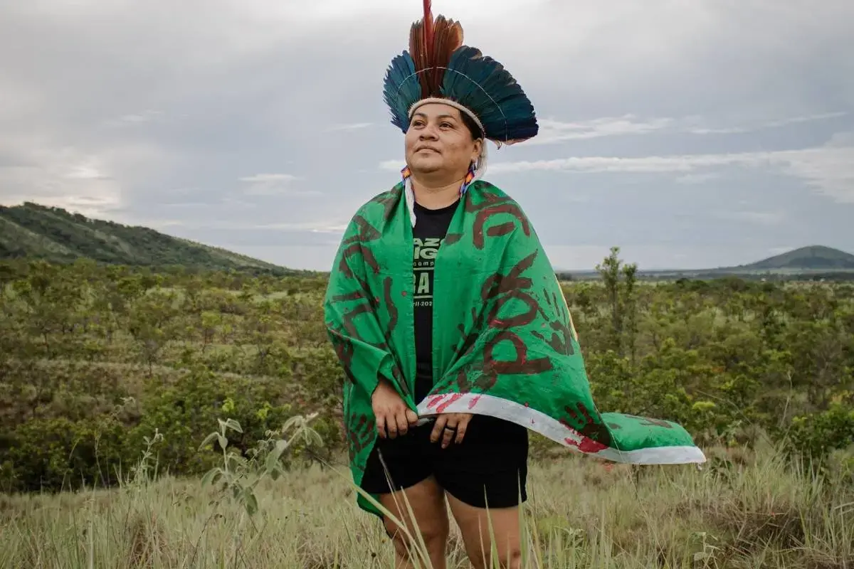 A woman wearing a blue and red headdress looks out at the horizon. she is standing high grass with mountains in the background. She is wearing a green flag with red writing and handprints around her shoulders.