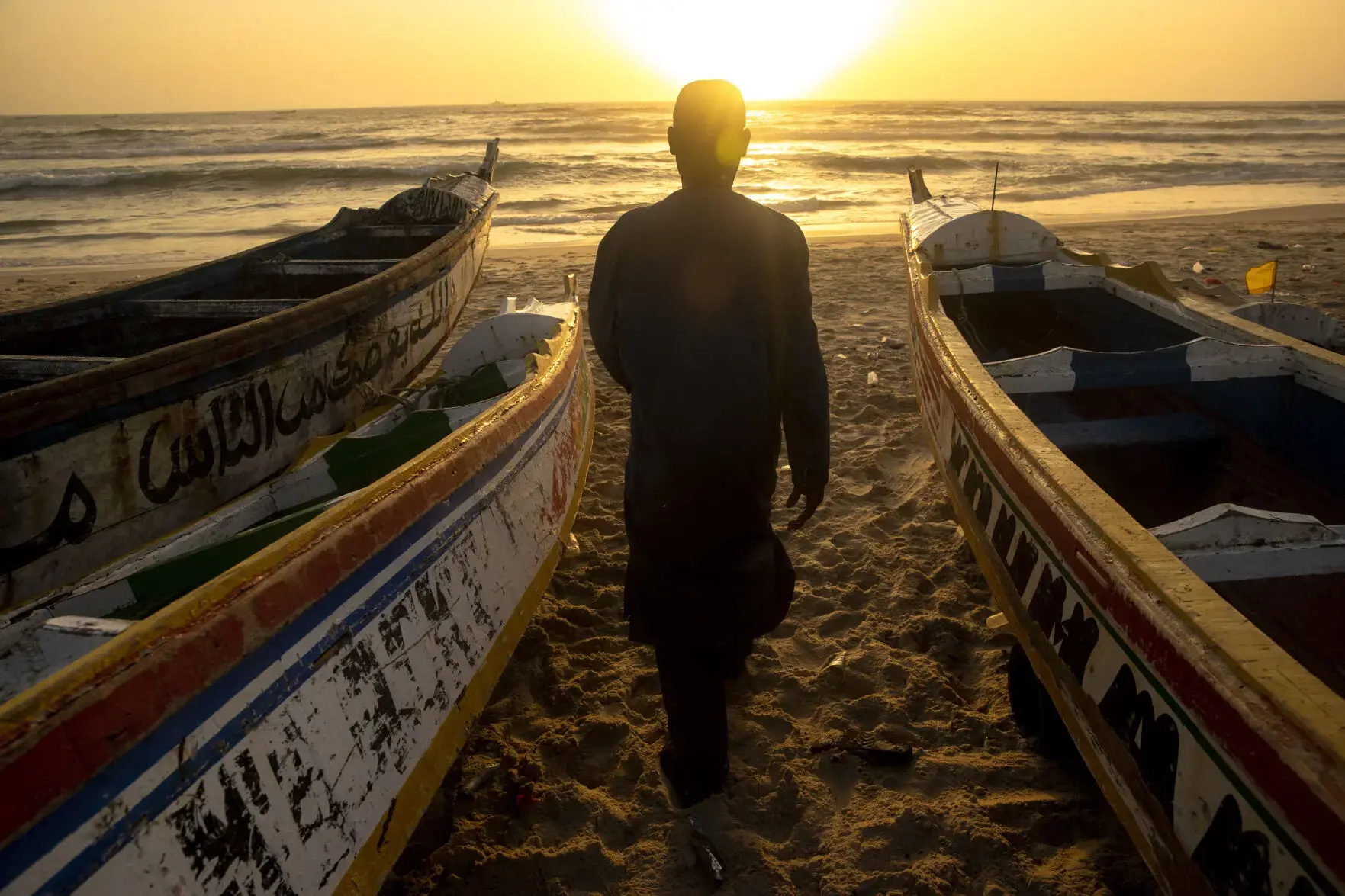 The back of a man's silhouette is visible at sunset, looking out at the ocean in between three wooden and colorful fishing boats.