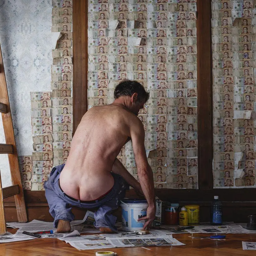 A man is seen shirtless painting pesos to the wall in his home
