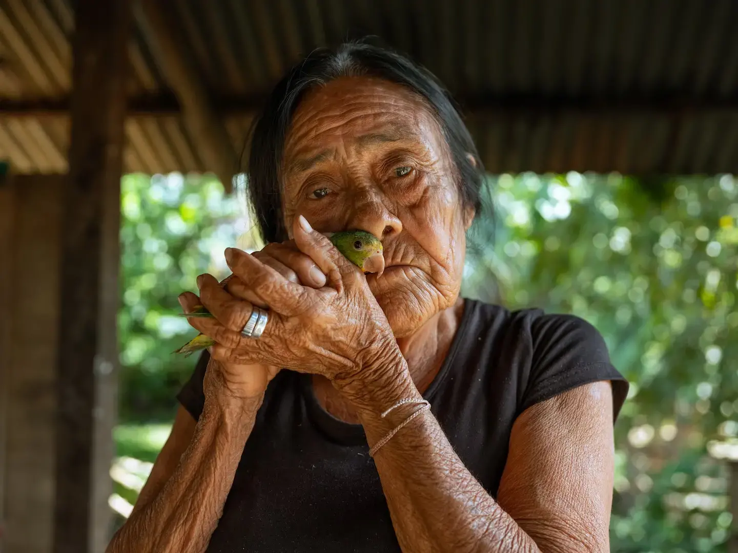 An elderly woman stands holding a green bird close to her face