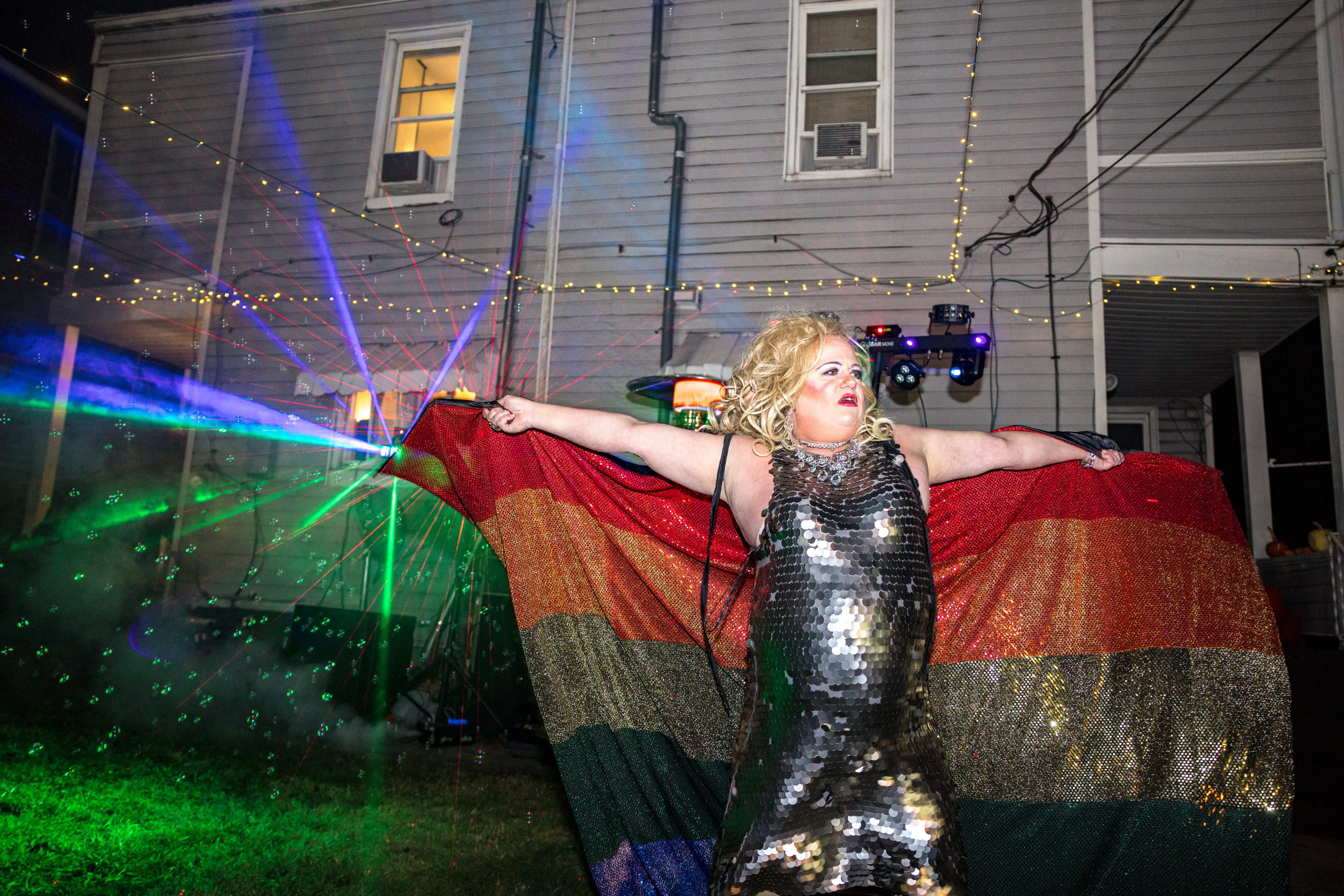 A drag queen poses wearing a sequin dress and holding a rainbow flag made of sequins.