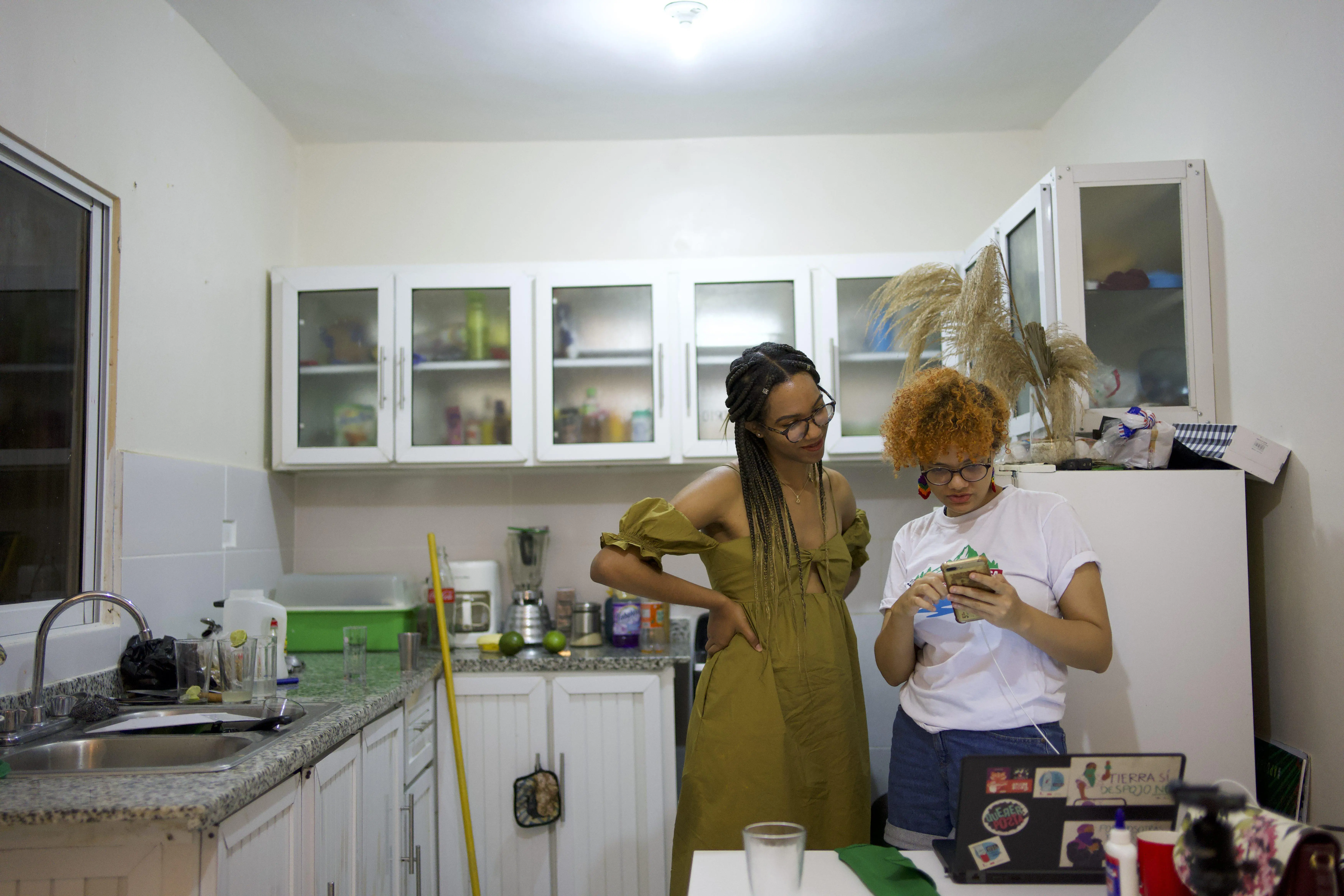 Two women stand in a kitchen while one shows the other women something on her phone.