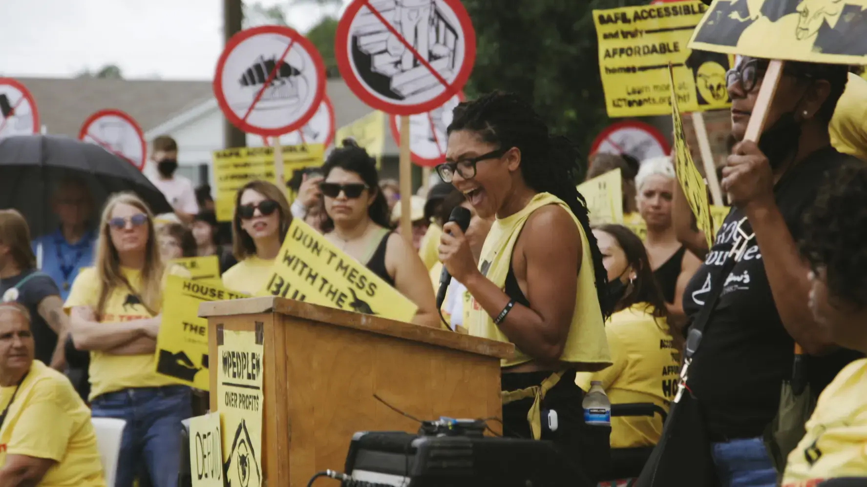 A woman in yellow speaks at a podium at a protest, surrounded by other protestors. The protestors are carrying yellow signs that say 