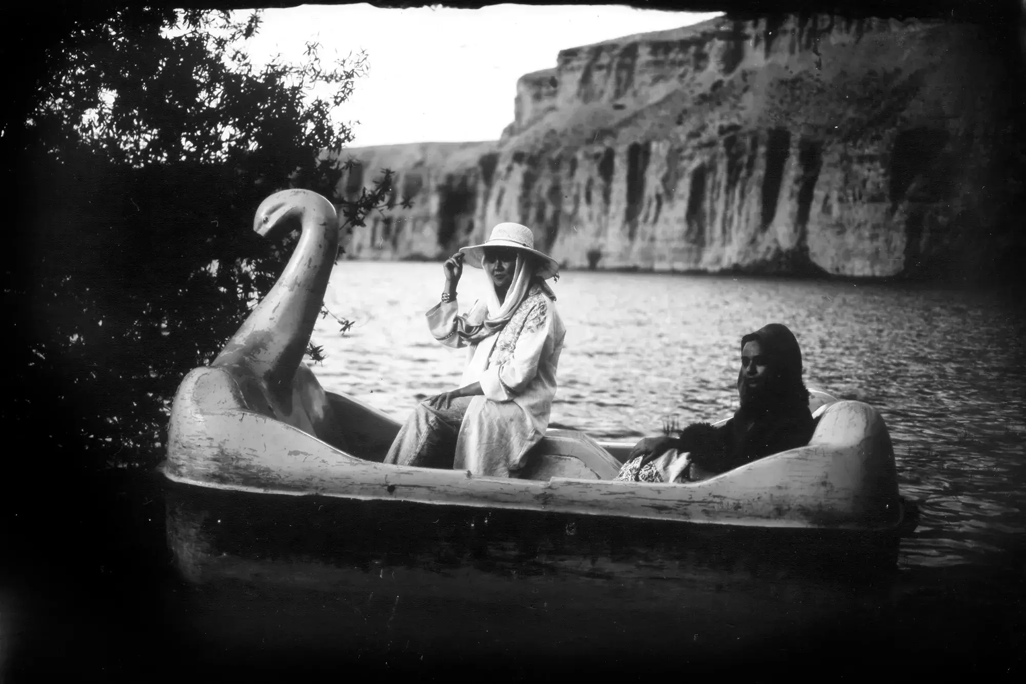 In a black and white photograph, two women sit in a swan boat. One woman is wearing a hat with a scarf. Cliffs are visible in the background.