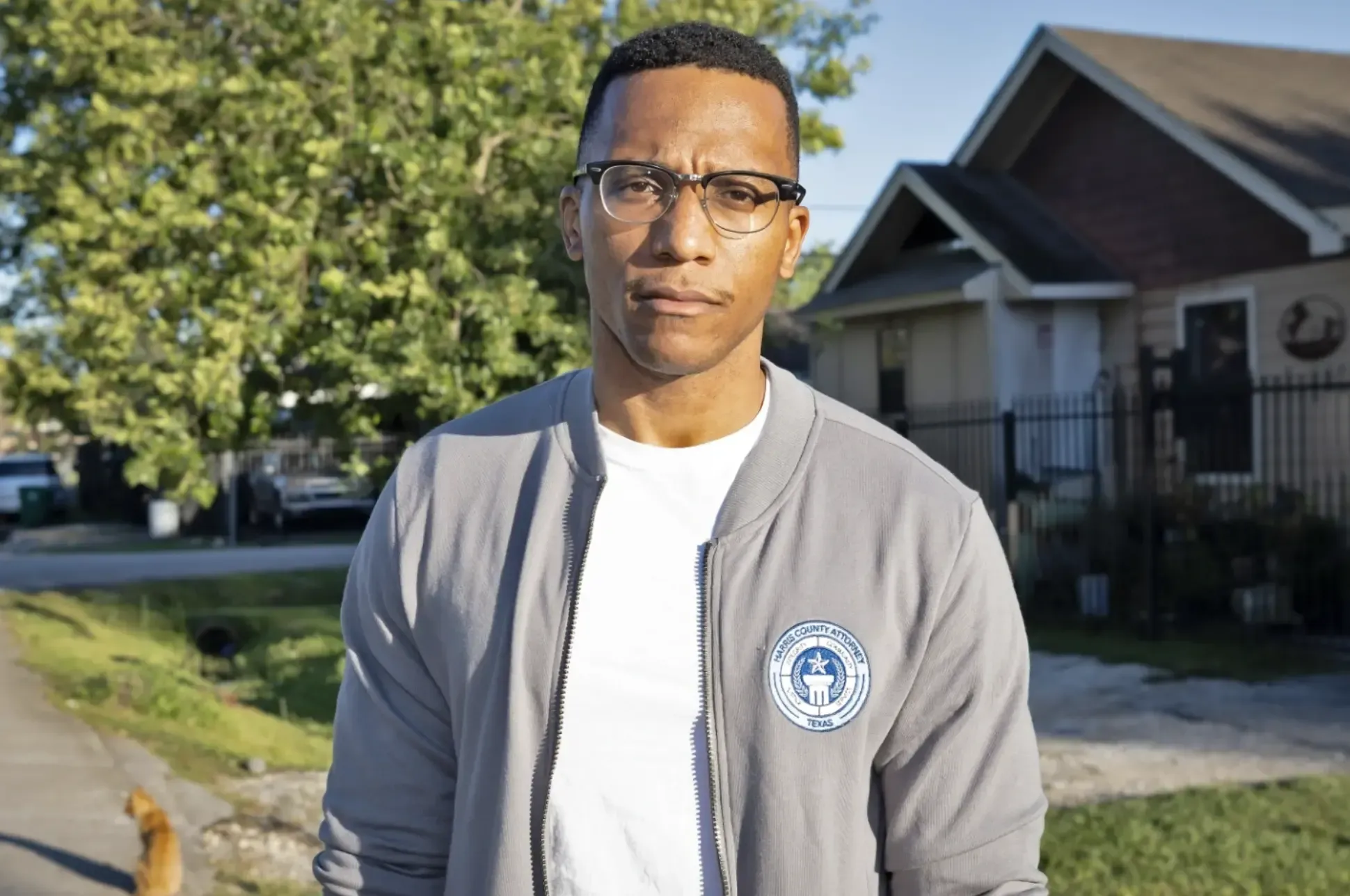 A man with glasses stands in county attorney grey bomber jacket in front of a home and a tree