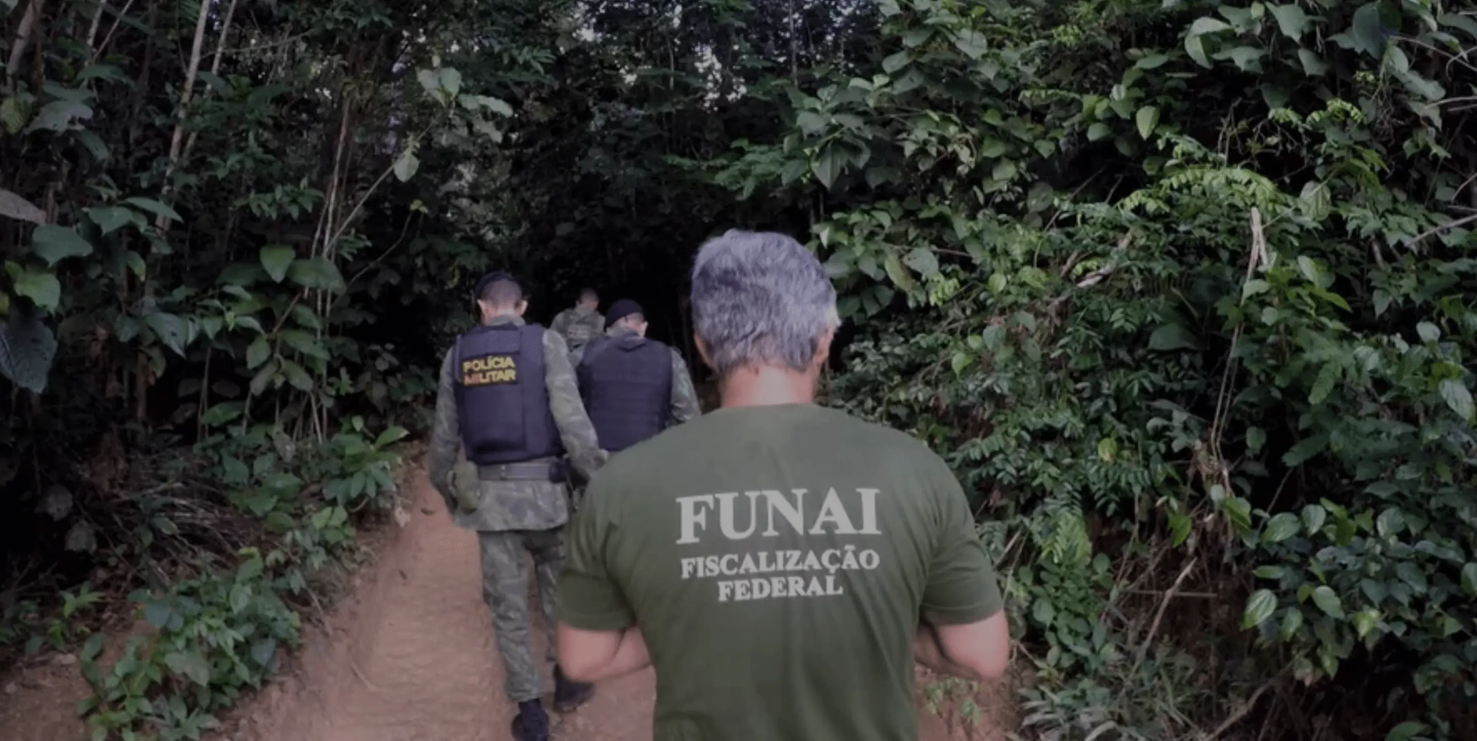 Men in uniform walk into the rainforest; one wears a FUNAI shirt while others are in police gear.