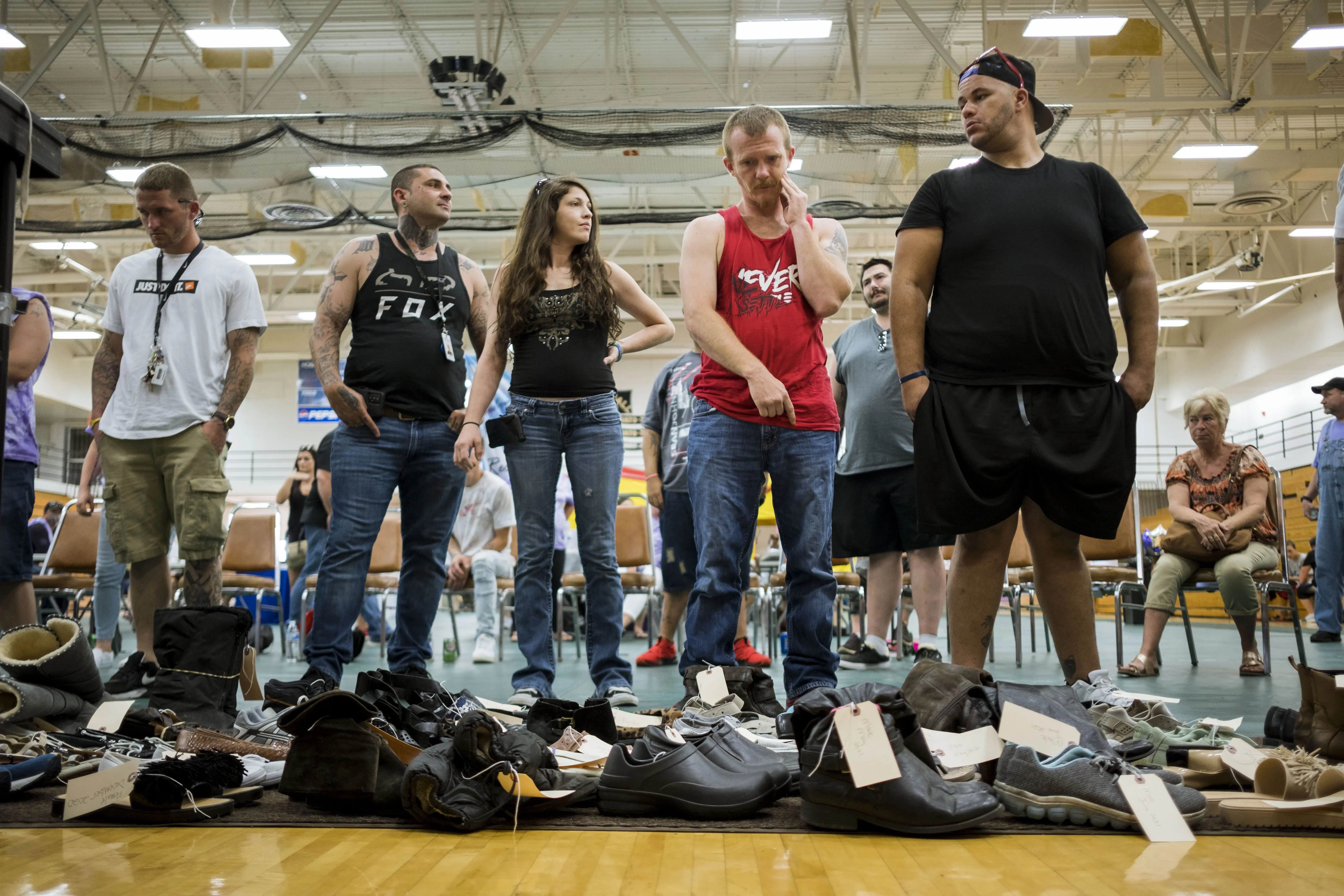 Five people line up inside of a gym. Shoes, each with a descriptive tag, sit empty before them.