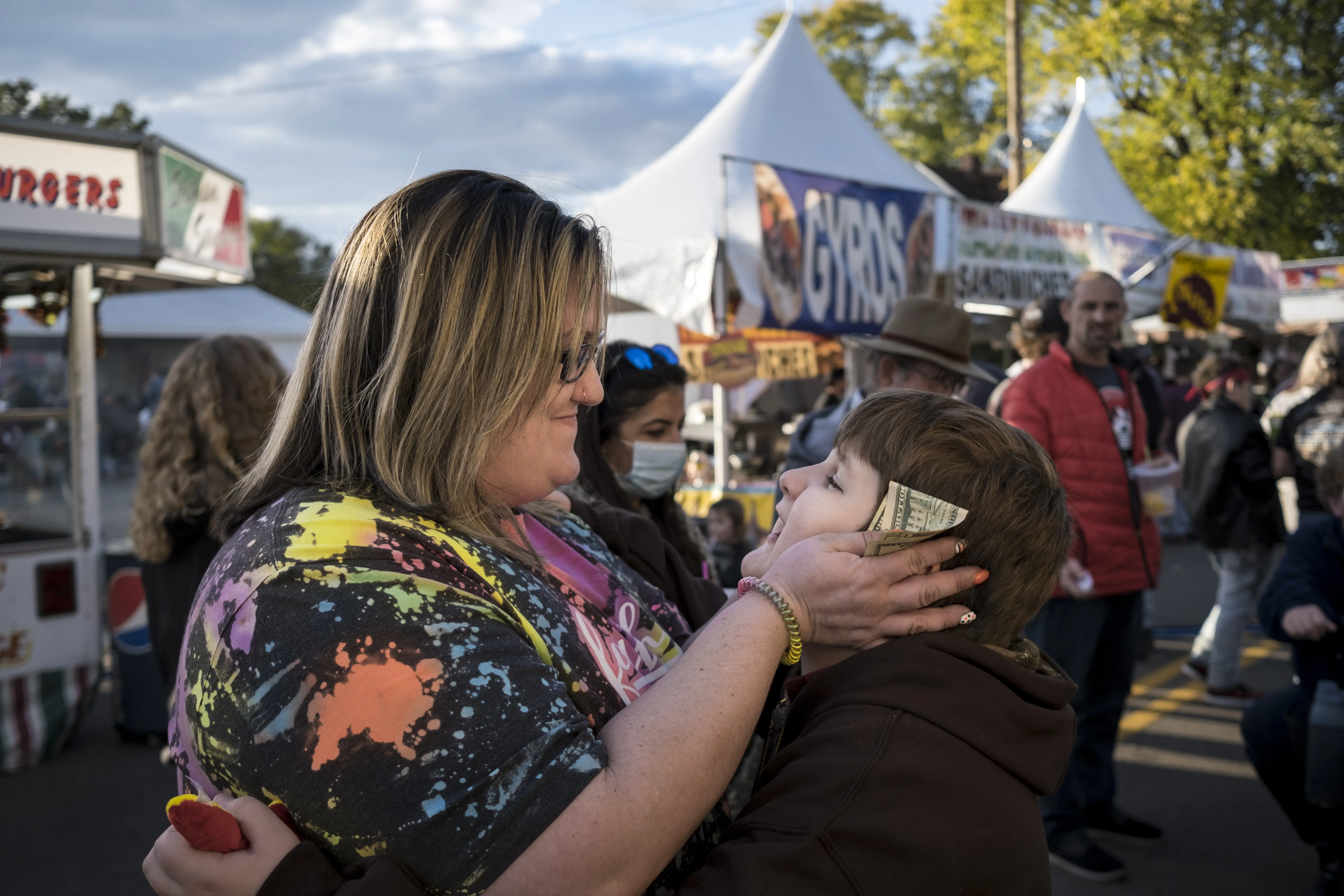 A woman, shown from the side in front of street vendors, frames a child's face in her hands. She has slipped a dollar bill between her thumb and pointer. The child holds her waist.