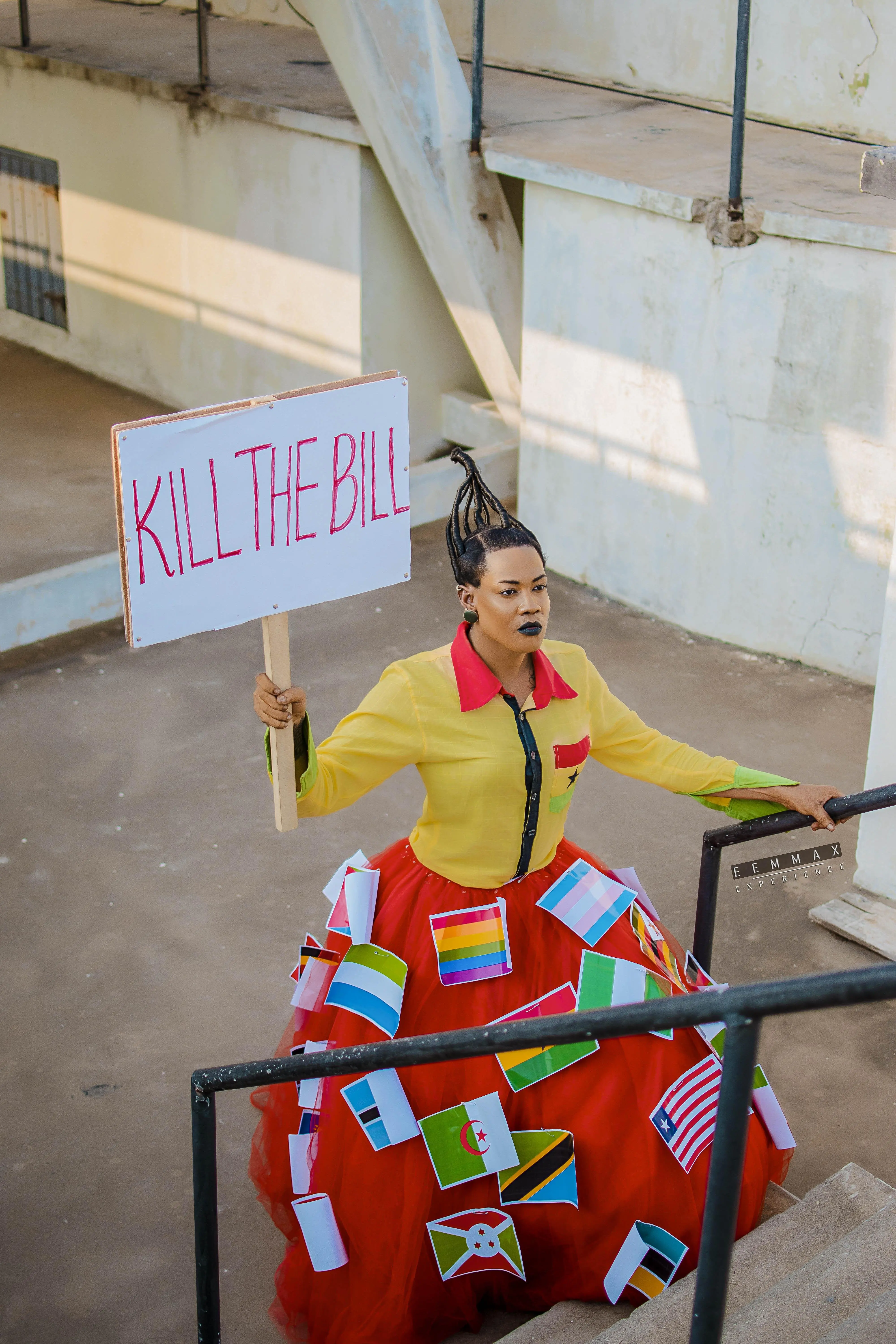 A transgender advocate holds a sign reading Kill the Bill, wearing a dress decorated with world flags.
