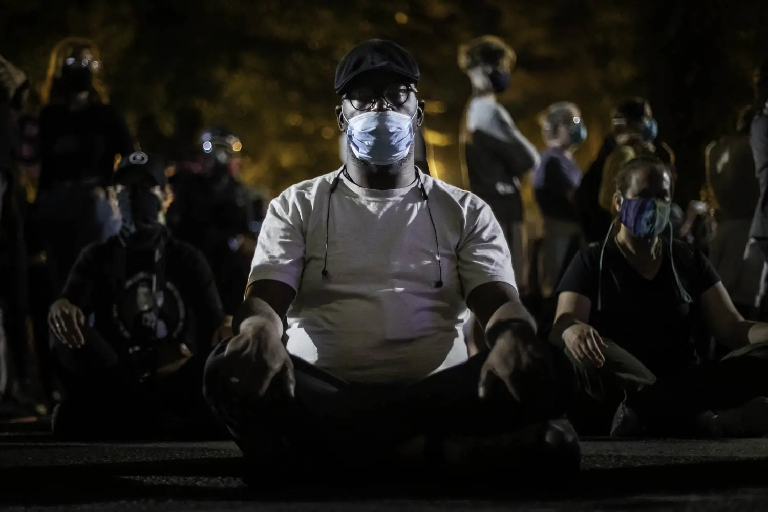 A man wearing a medical face mask sits on the ground alongside others.