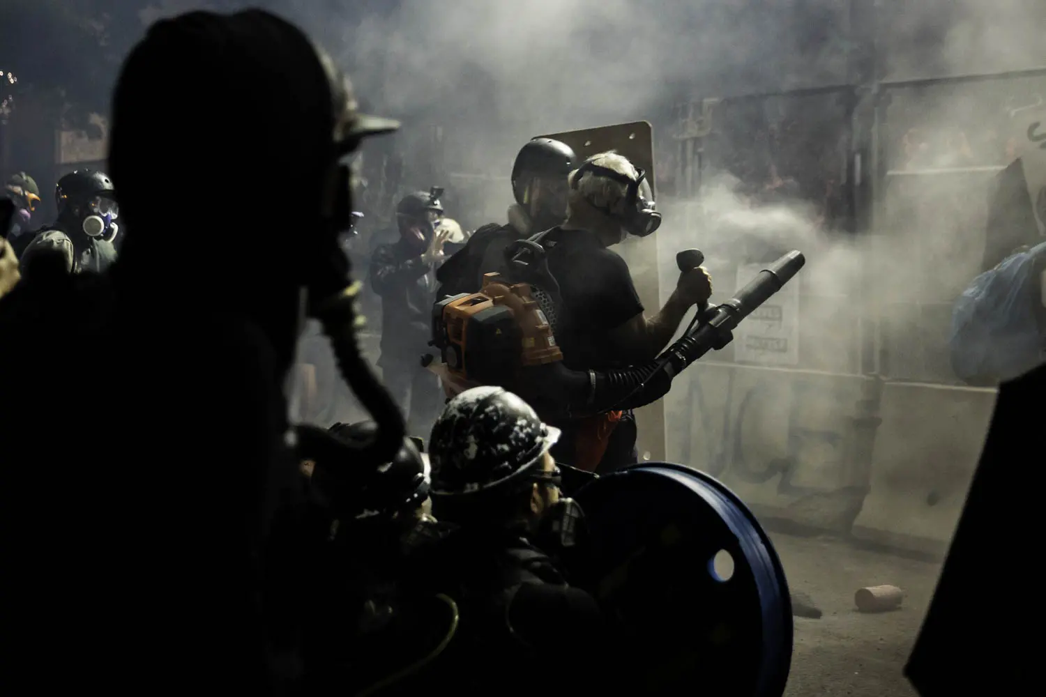 A man in a gas mask and with a leafblower stands next to a police barricade as tear gas fills the air