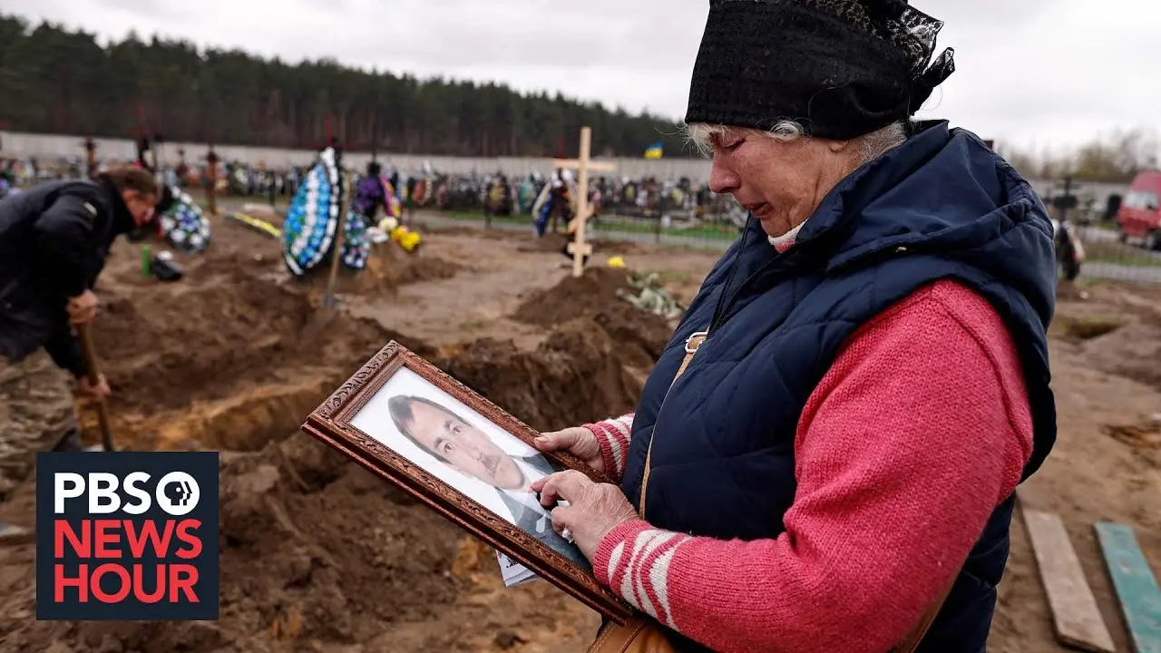 A woman holds a photo of a man while holding a tissue in a graveyard.