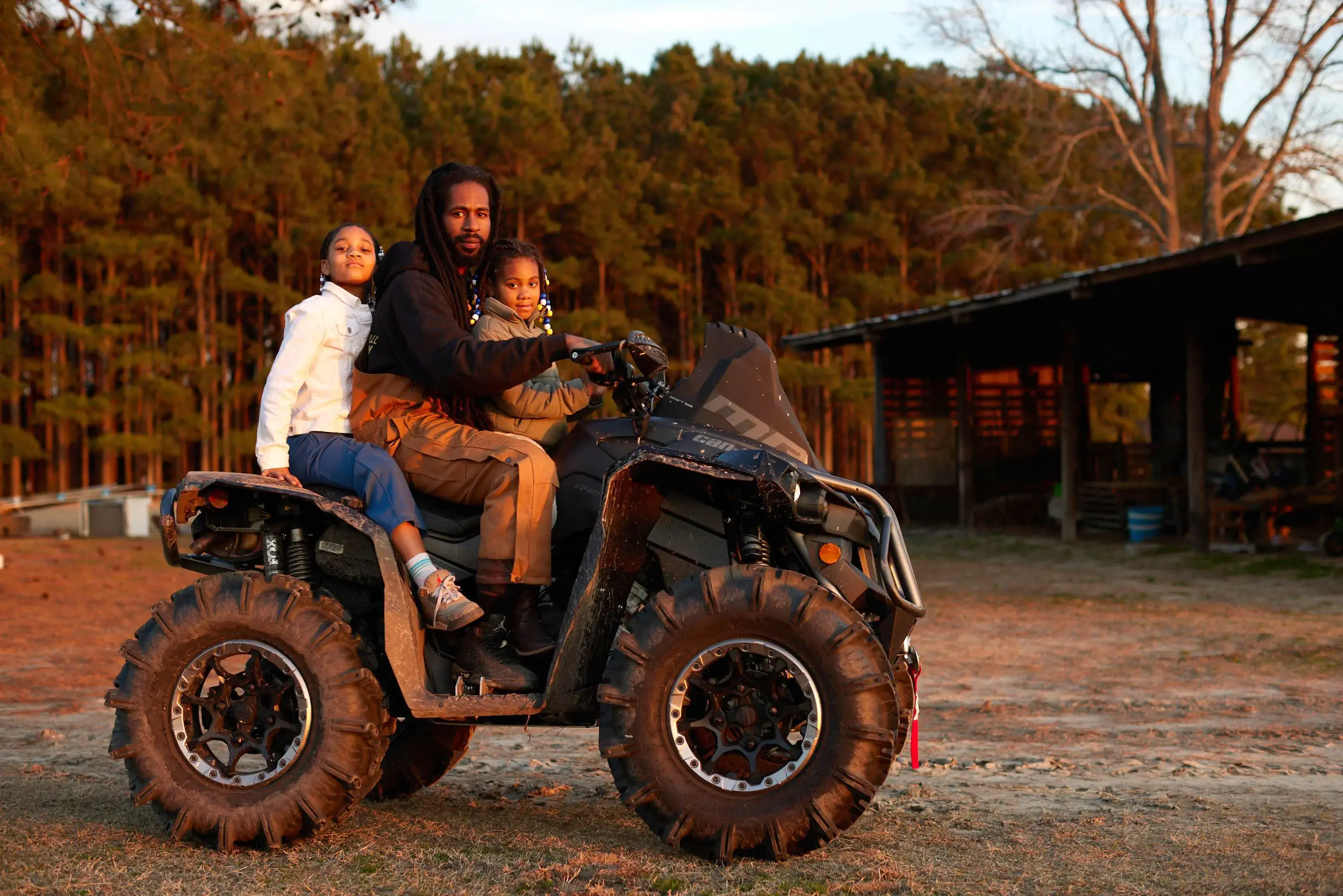A man sits on a tractor between his two young daughters during sunset.