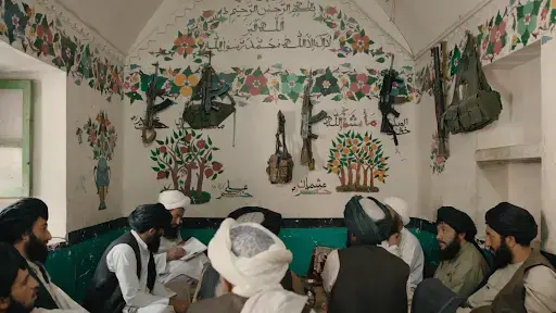 Men crowd in a small room with flowers pained on white walls. They are sitting on a green couch and actively engaged in conversation. Guns and ammunition hang from the walls above the men's heads.