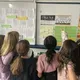 group of young girls face the whiteboard and point to documents taped to the board